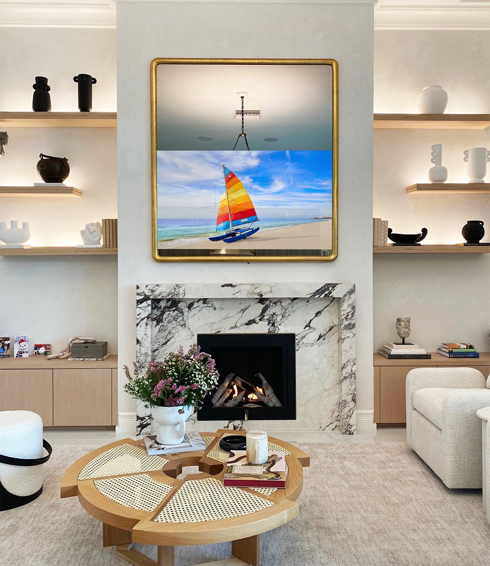 round corner vertical gold Mirror TV frame, multi colored catamaran on TV, in staged Living Room over marble fireplace, beige chairs and rug