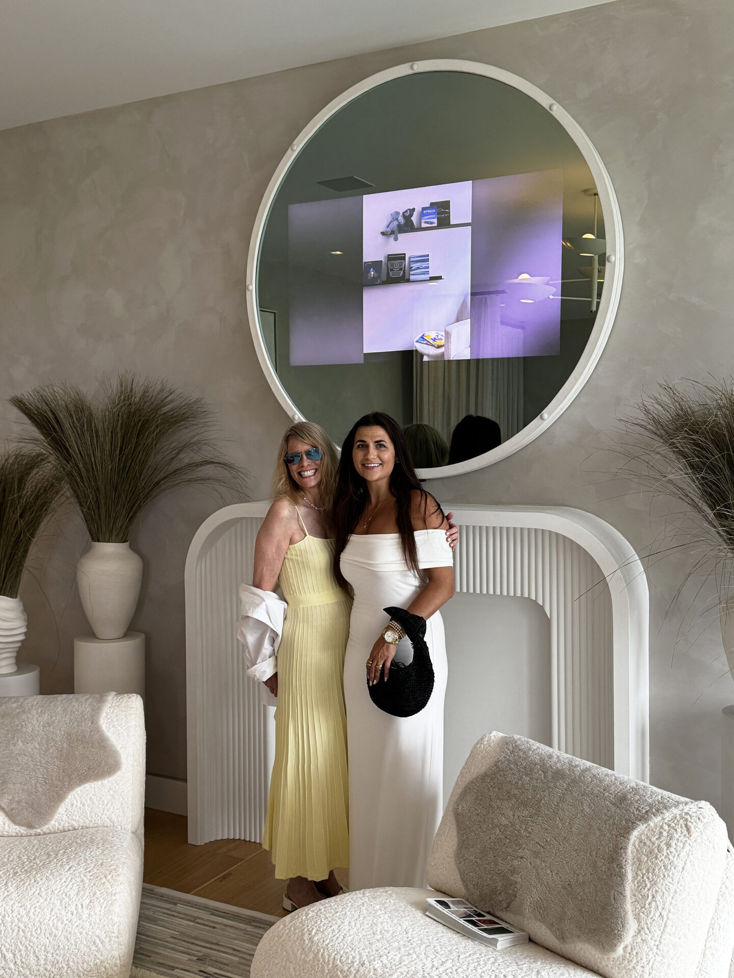 Reflectel Owner Elizabeth Goldfedder Posing In Front White Leather Circle Framed Mirror Television Next To Designer Brianna Scott In Living Room Showhouse