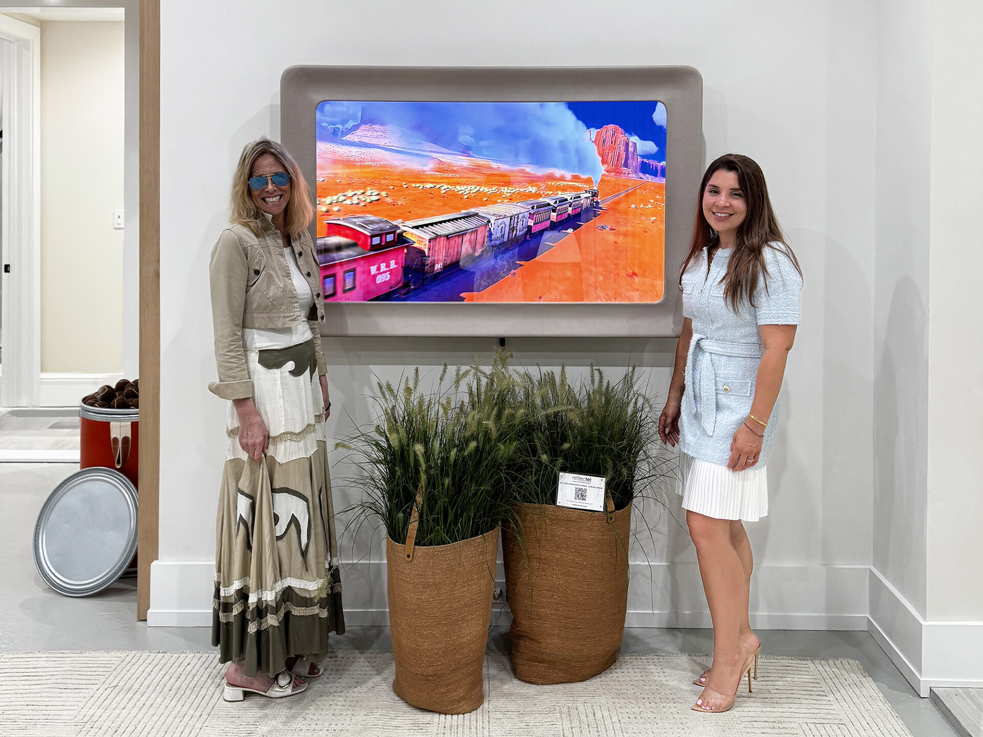 Reflectel Owner Elizabeth Goldfedder Posing In Front Of A Swoop Grey Leather Framed Mirror Television Next To Designer Daniella Feldman In Living Room Showhouse