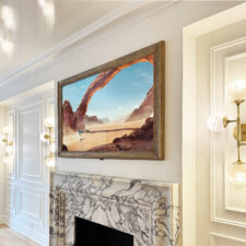 65" Recessed Landscape - Gilded Carved textured golden brown Reflectel mirror television turned on showing a canyon scene above marble mantel inside living room
