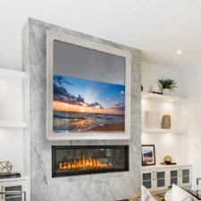 65" Rounded Corner "Swoop" - Cortina Leathers Suede White suede lather square rounded corner mirror television turned on displaying image of a sunset beach above a marble mantel in living room